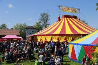 Das Bild zeigt bunte Zirkuszelte und viele Menschen auf einer Wiese.