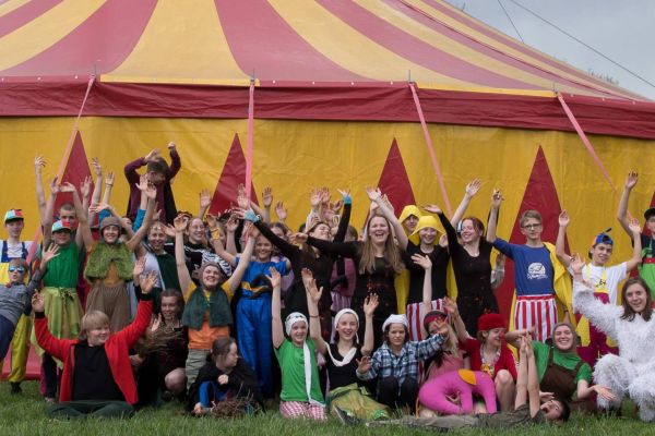 Kinder und Jugendliche posieren in Kostümen vor einem Zirkuszelt und jubeln.