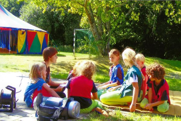 Sechs Kinder sitzen mit ihrer Trainerin auf einer Wiese vor einem kleinen Zirkuszelt und die Sonne scheint.