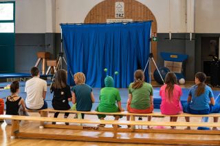 Junge Zirkusartis*innen warten vor der aufgebauten Manege sitzend auf Holzbänken auf ihren Auftritt.